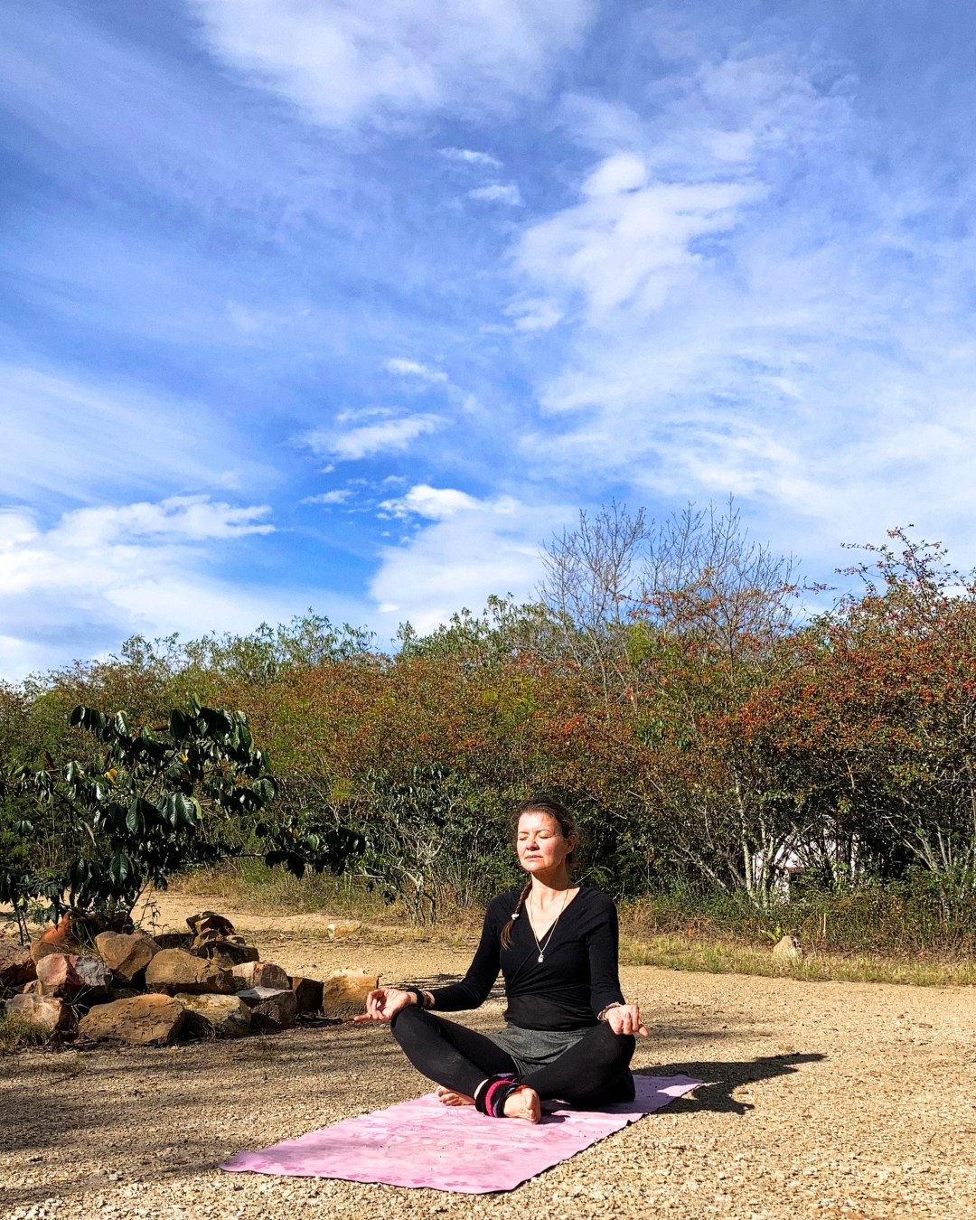 Simone im Lotussitz in Meditation unter weitem blauen Himmel, umgeben von Sträuchern in der Natur